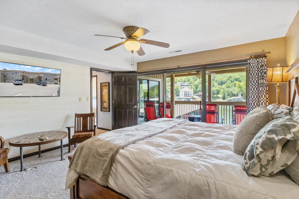 A comfortable bedroom with a plush bed and soft linens, large windows framing a breathtaking waterfront view, and a mounted TV for evening relaxation. A wooden chair and table by the window invite moments of reading or quiet contemplation.