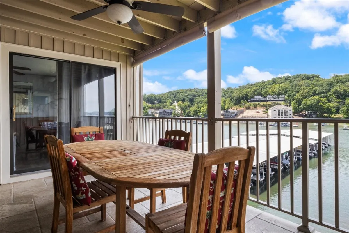 An inviting balcony with a wooden dining table and chairs overlooks the sparkling lake. The serene, open space is ideal for outdoor dining or sunset toasts, with a sense of connection to nature and scenic views.