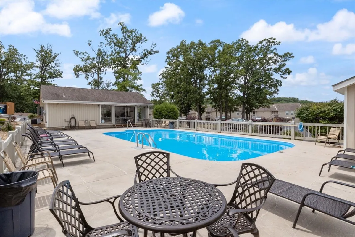 This image displays a sparkling outdoor swimming pool surrounded by plenty of seating and lush trees. The adjacent clubhouse is visible, suggesting easy access to indoor amenities, while the inviting poolside area implies a space ideal for relaxation and social gatherings.
