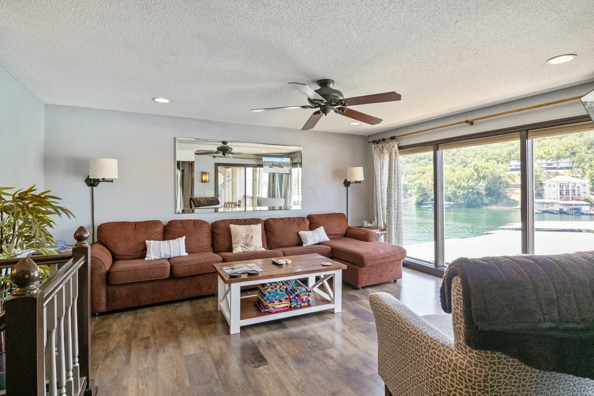 A bright living area with a large sectional sofa and coffee table, facing expansive windows that offer unobstructed lake views. The welcoming space is arranged for both relaxation and socializing, with natural light spilling in and a sense of connection to the outdoors.