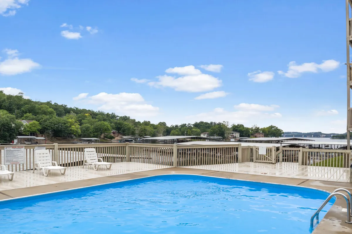 The hero image showcases the exterior of the property, with an inviting swimming pool surrounded by comfortable lounge chairs. Beyond the pool, there's a stunning waterfront view of the serene cove and main channel, perfect for watching sunsets and enjoying the natural beauty of Osage Beach. The setup suggests opportunities for sunbathing, swimming, and gathering with friends or family in a relaxing, picturesque setting.