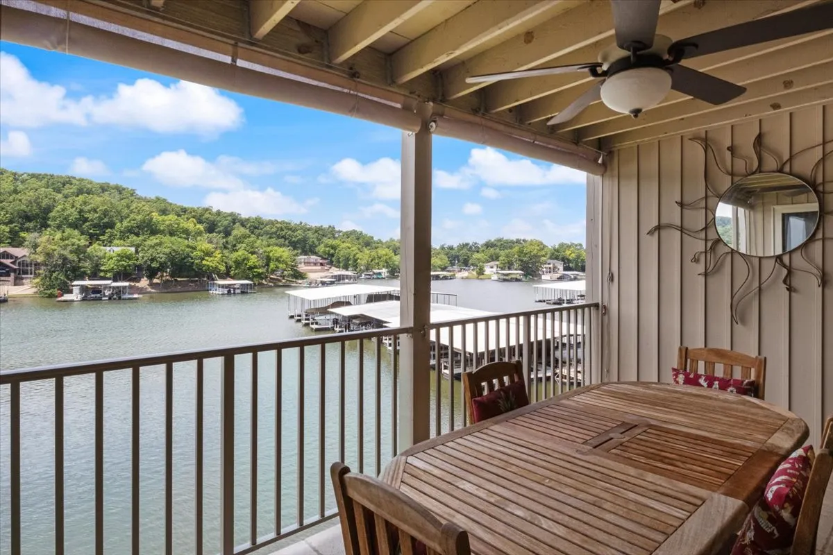 An exterior lakeside dining area with an outdoor table, surrounded by green trees, and a clear view of the water and boathouses. The scene is serene, ideal for alfresco meals or relaxing by the water’s edge.