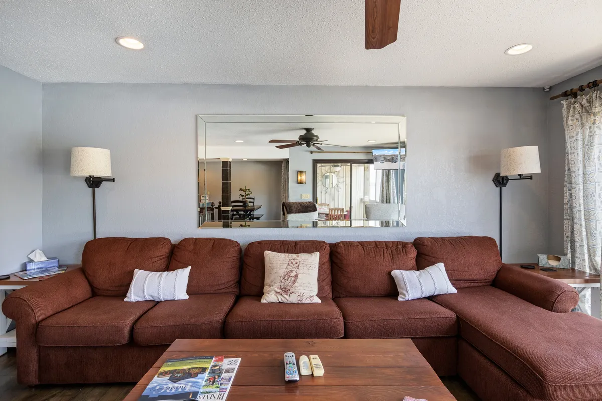 A large brown sectional sits beside a coffee table adorned with magazines and remotes. The mirror in the background reflects a view of the dining area, creating a sense of openness and togetherness.