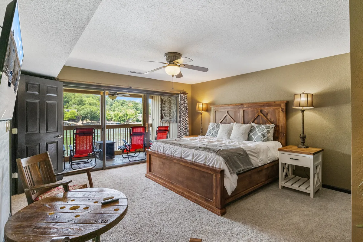 A spacious bedroom featuring a king-size bed adorned with decorative pillows faces a balcony furnished with vibrant red chairs. The room is bathed in natural light from large windows, and a wooden side table completes the cozy, welcoming atmosphere. The balcony offers a direct view of the lake beyond.