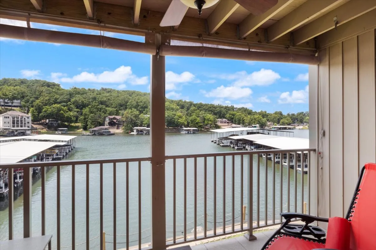 A tranquil lake view from a balcony, with boathouses dotting the shoreline and green hills beyond. The balcony railing suggests a private vantage point for guests to unwind while taking in serene natural surroundings.