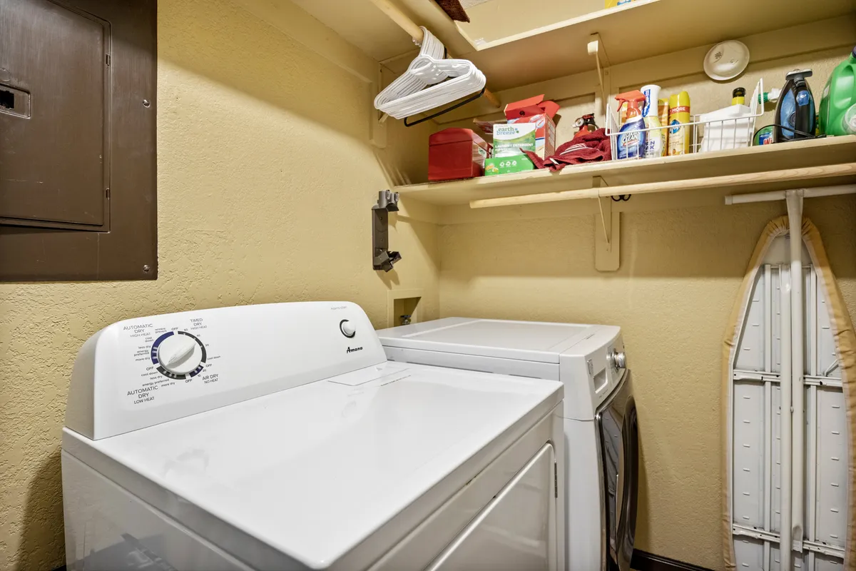 A tidy laundry area featuring a washing machine, dryer, shelves with supplies, and an ironing board. Everything is neatly organized and ready for guest use, supporting an effortless, fully prepared stay.
