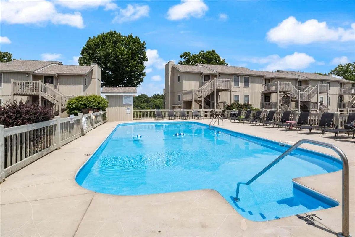 A sunlit community swimming pool is surrounded by comfortable lounge chairs and bordered by lush, mature trees. The inviting water sparkles under clear skies, while the seating area offers ample space for families and friends to relax poolside.