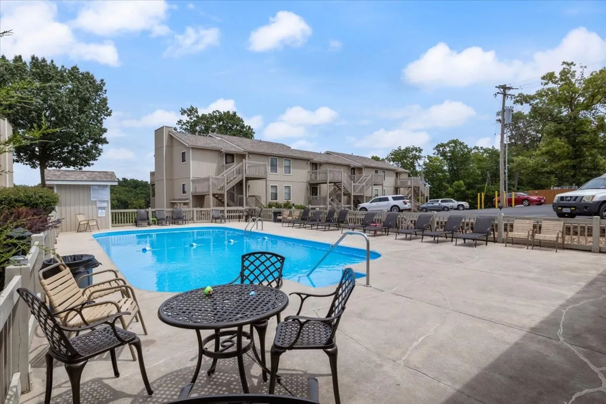 A bright, leisurely pool scene with ample outdoor seating, mature trees for shade, and a residential building in the background. The pool area feels both private and communal, encouraging social interaction and relaxation.